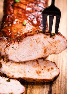 A cutting board with a BBQ pork loin and a few slices in the front, and a fork picking one up.