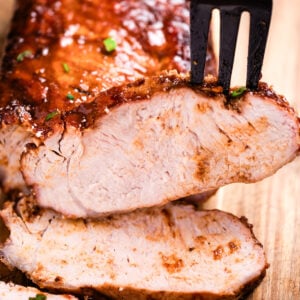 A cutting board with a BBQ pork loin and a few slices in the front, and a fork picking one up.