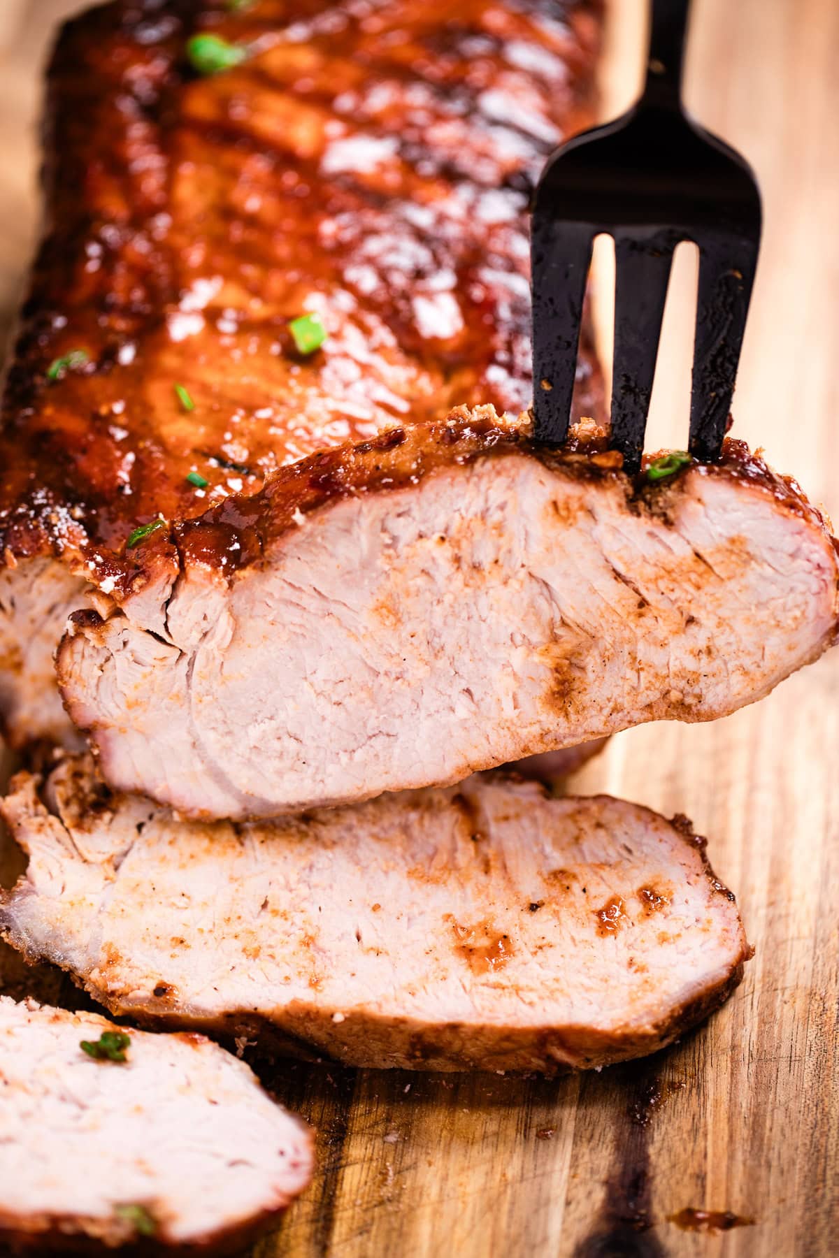 A cutting board with a BBQ pork loin and a few slices in the front, and a fork picking one up.