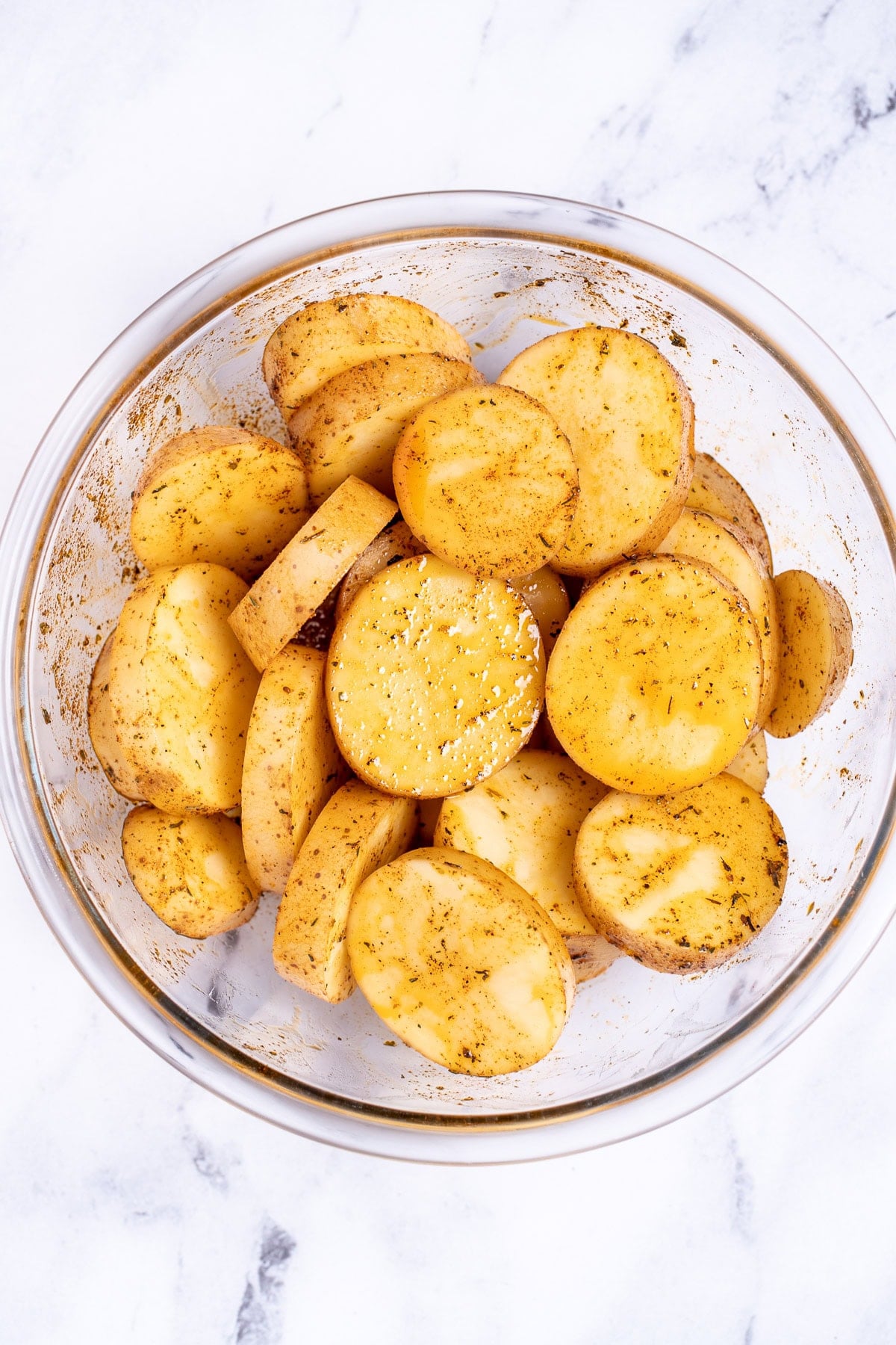 A mixing bowl on a table, with potato slices tossed with olive oil and seasoning.