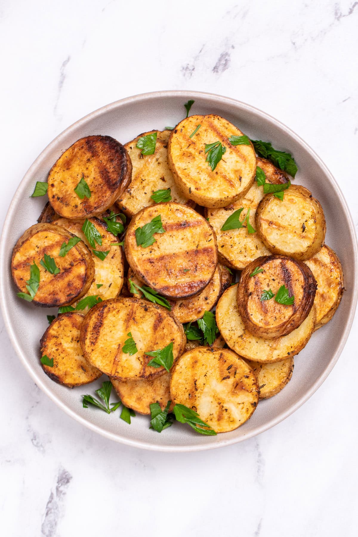 A serving bowl with grilled potato slices topped with chopped fresh parsley. The potato slices have nice grill marks.