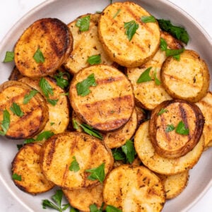 A serving bowl with grilled potato slices topped with chopped fresh parsley. The potato slices have nice grill marks.