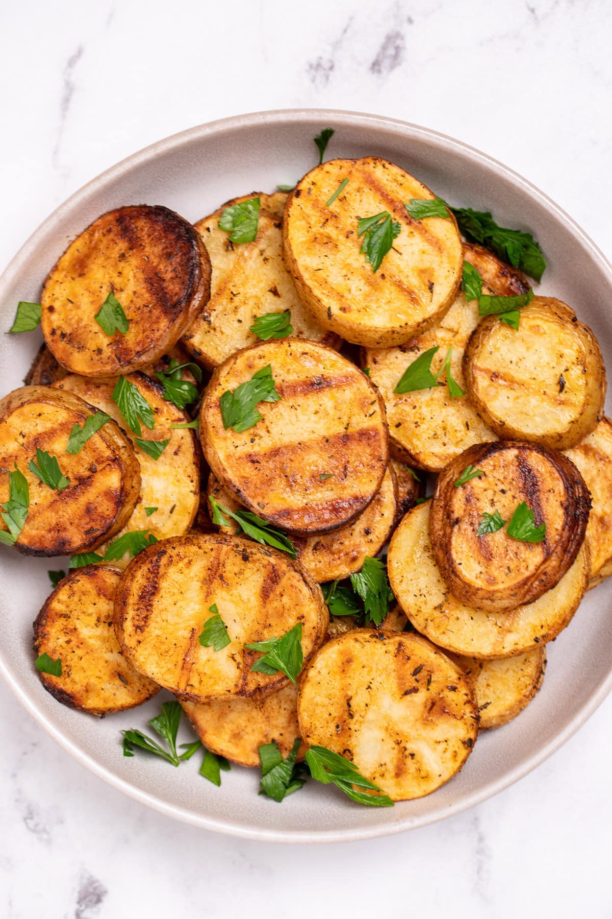 A serving bowl with grilled potato slices topped with chopped fresh parsley. The potato slices have nice grill marks.