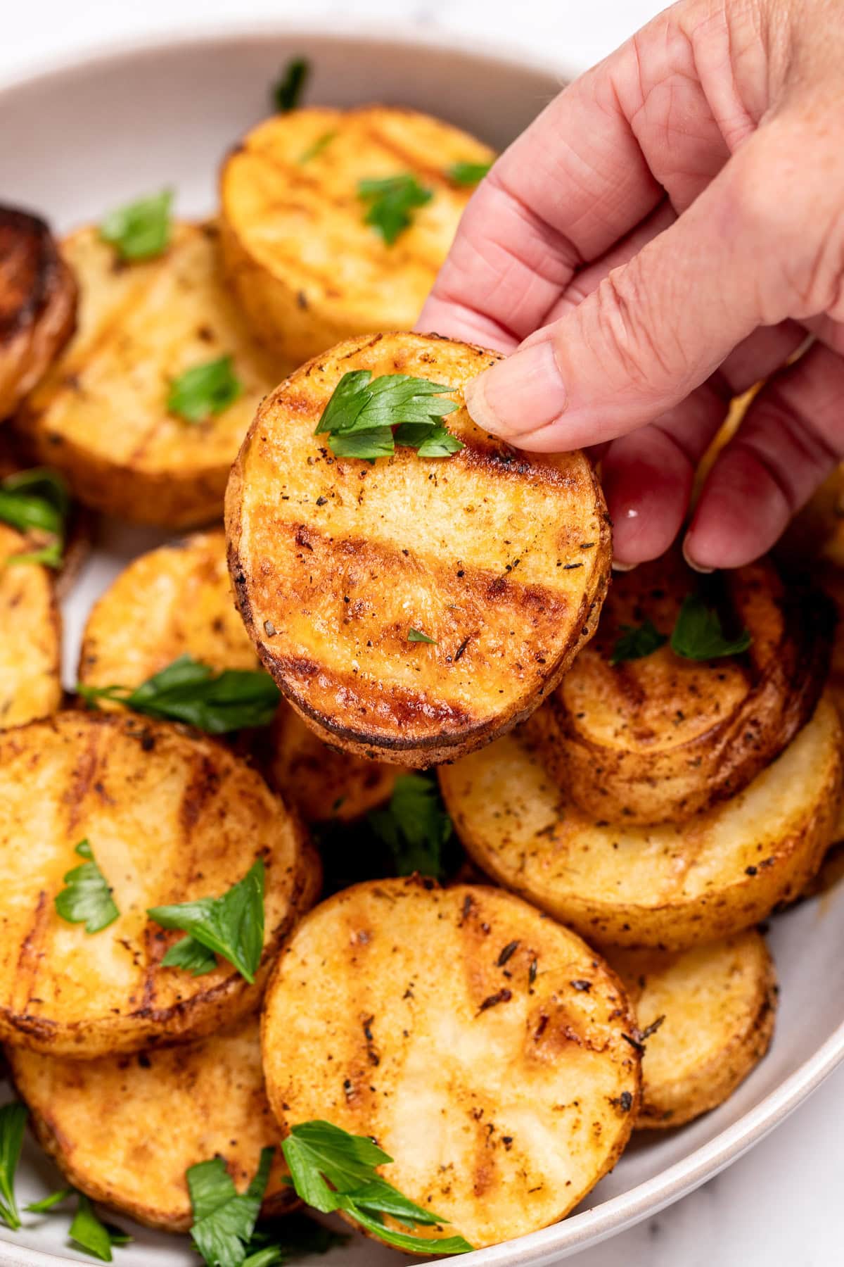 A serving bowl with grilled potato slices topped with chopped fresh parsley, and a hand holding one up. The potato slices have nice grill marks.