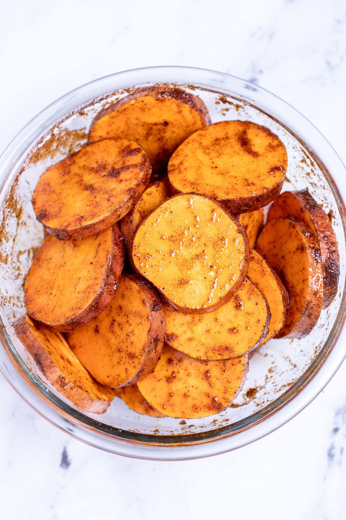 A bowl on a table, with sweet potato slices tossed with seasoning before being grilled.