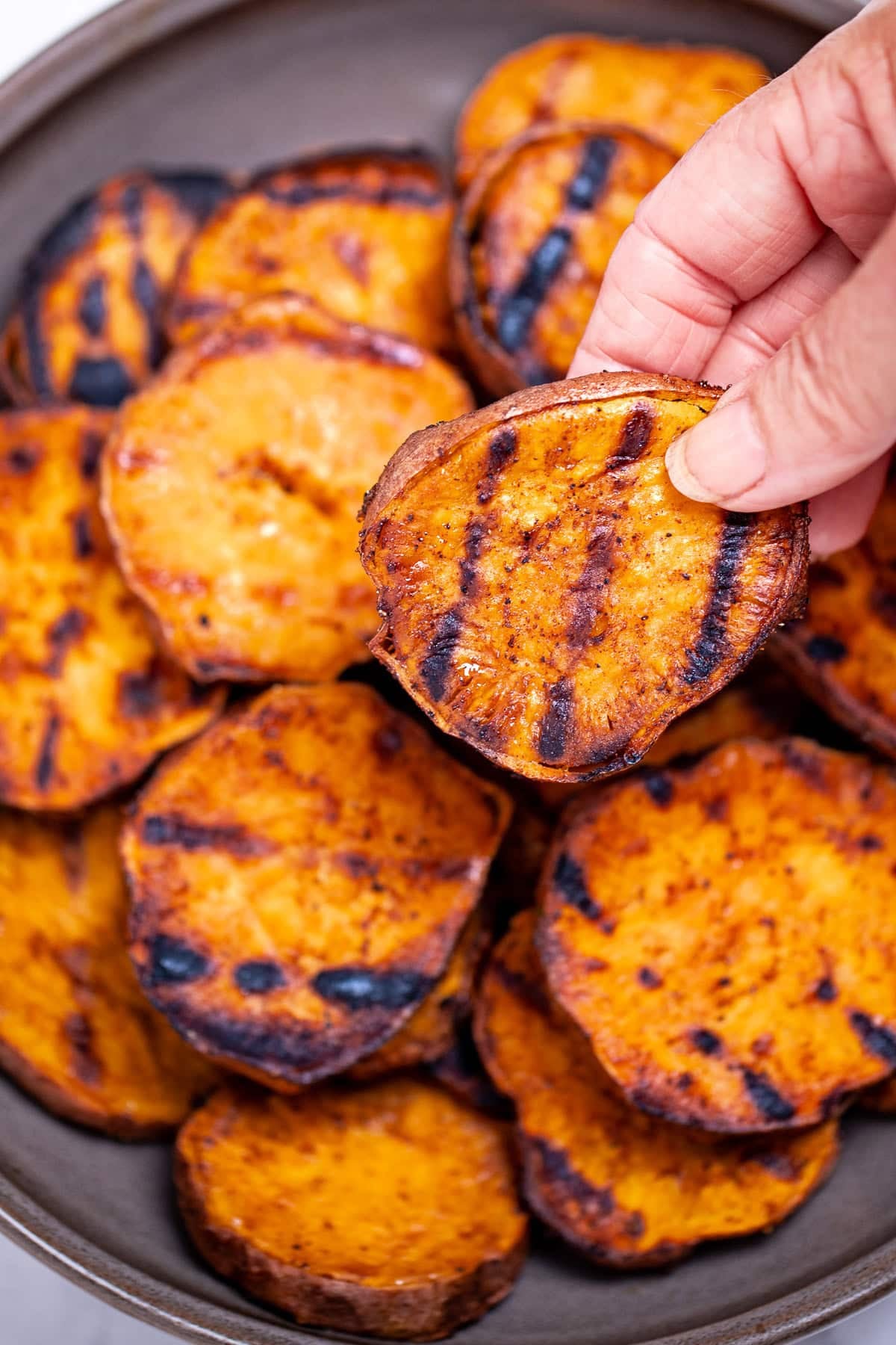 A plate of grilled sweet potatoes on a table, with a hand lifting one up.