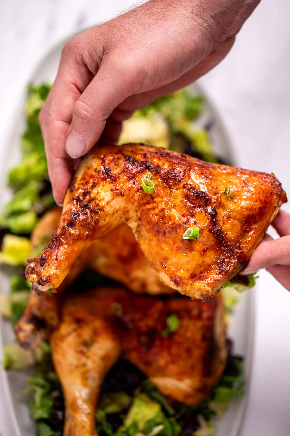 Three chicken quarters on a serving dish on top of salad greens, with two hands lifting one up.