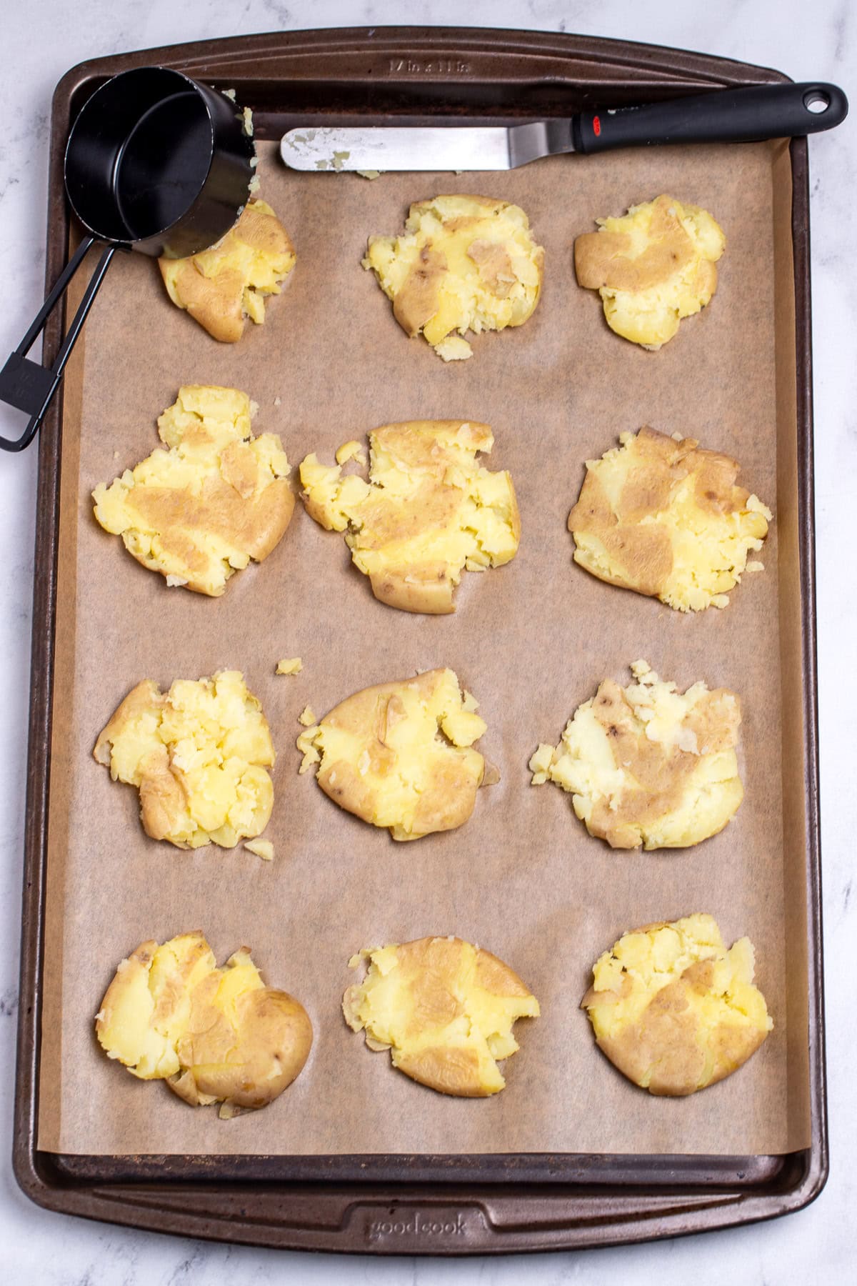 A sheet pan lined with parchment paper, with smashed potatoes next to a measuring cup and a spatula.
