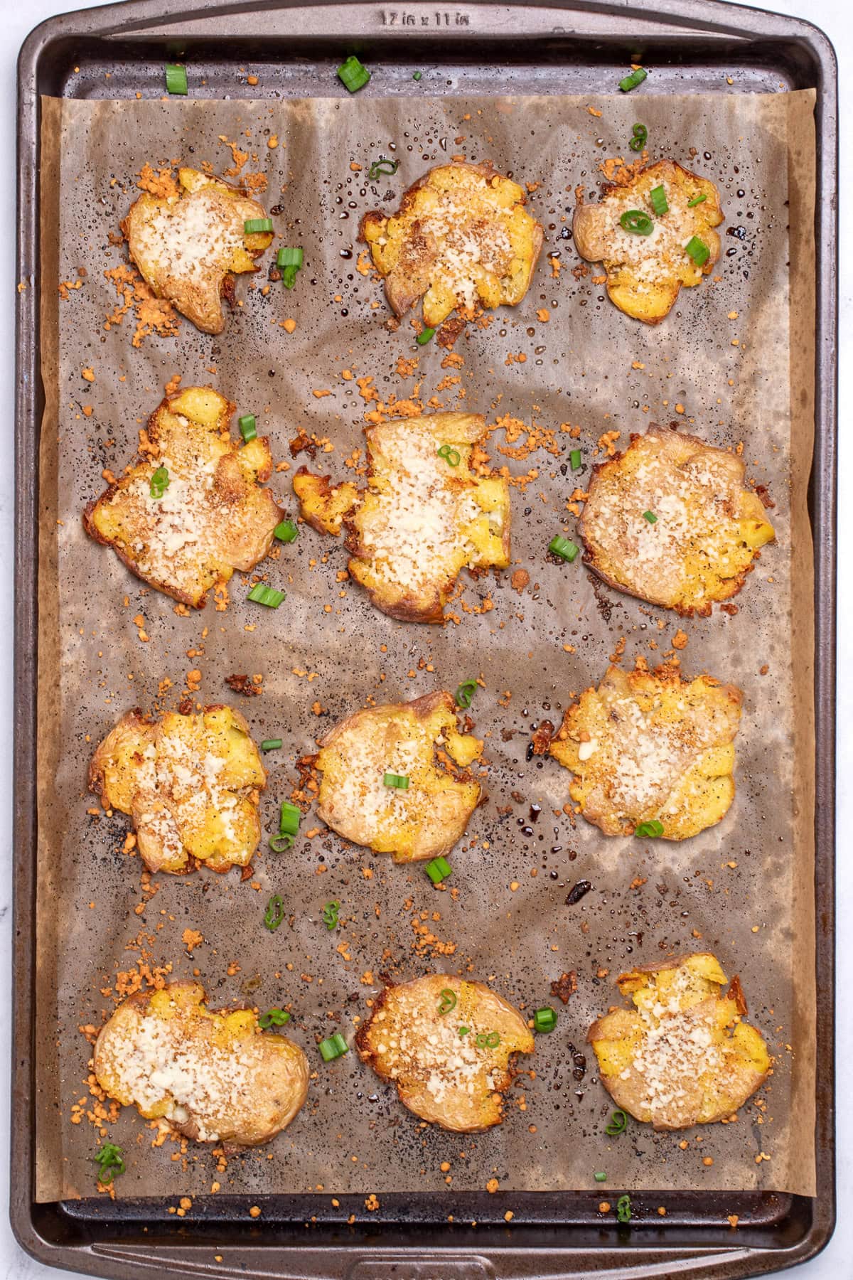 A sheet pan lined with parchment paper, with crispy roasted smashed potatoes topped with parmesan cheese and green onion.