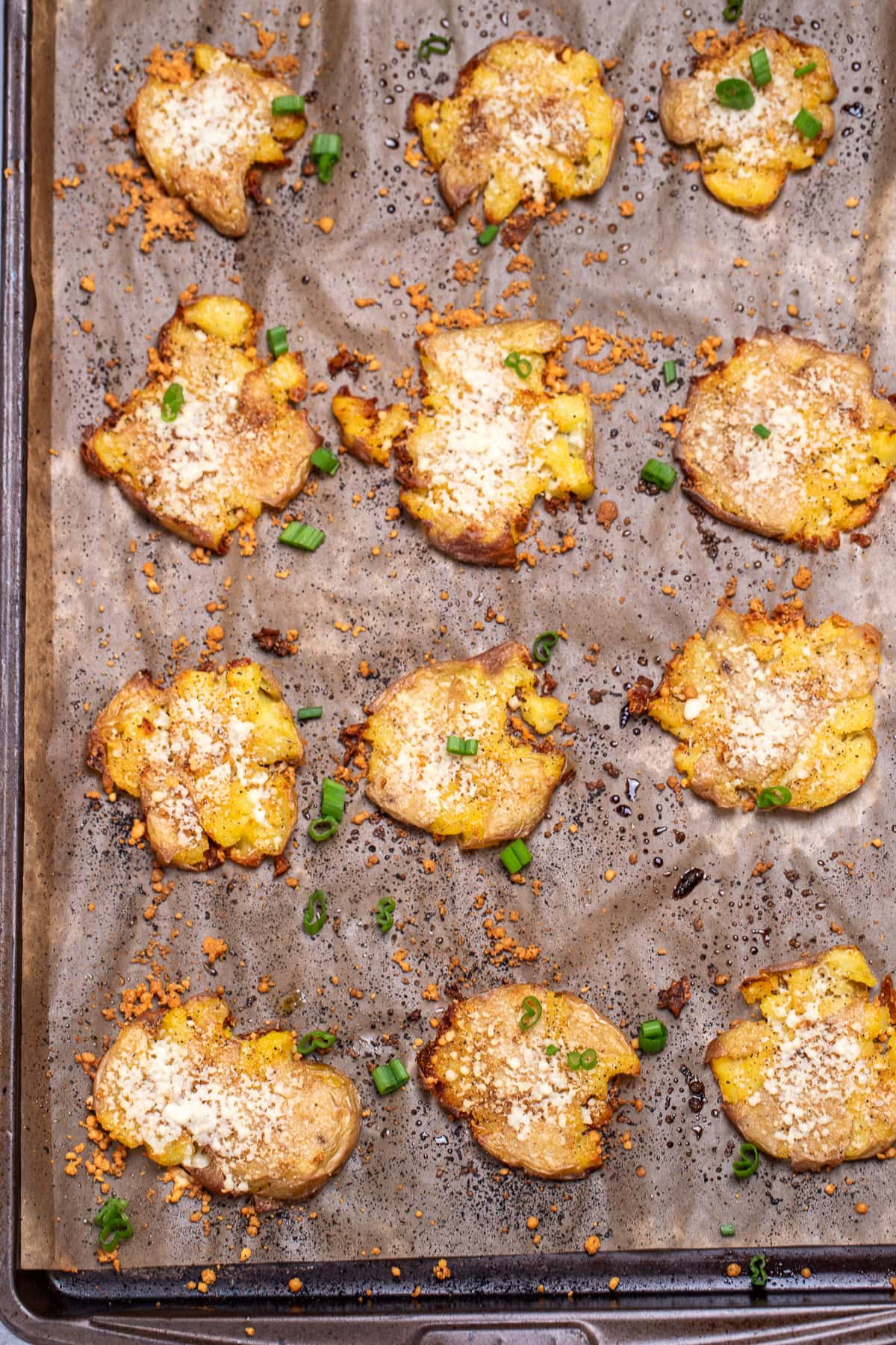 A sheet pan lined with parchment paper, with crispy roasted smashed potatoes topped with parmesan cheese and green onion.