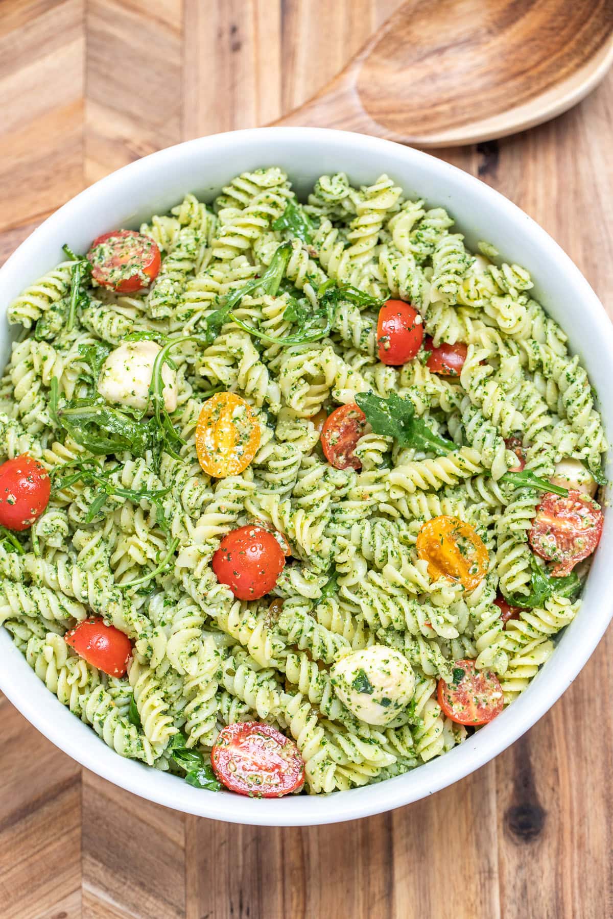 Pesto pasta salad with mozzarella and cherry tomatoes in a serving bowl resting on a cutting board.