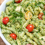 Pesto pasta salad with mozzarella and cherry tomatoes in a serving bowl resting on a cutting board.