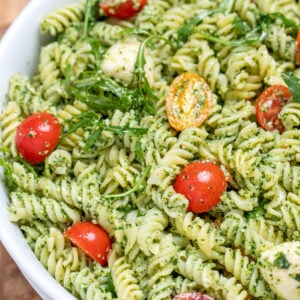 Pesto pasta salad with mozzarella and cherry tomatoes in a serving bowl resting on a cutting board.