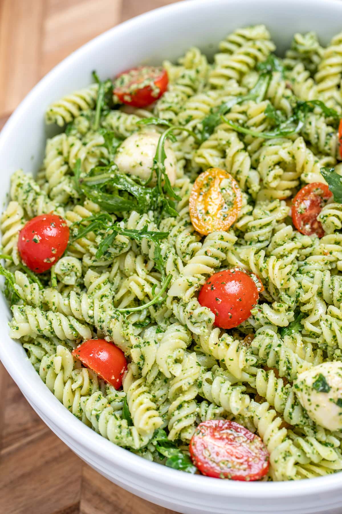 Pesto pasta salad with mozzarella and cherry tomatoes in a serving bowl resting on a cutting board.