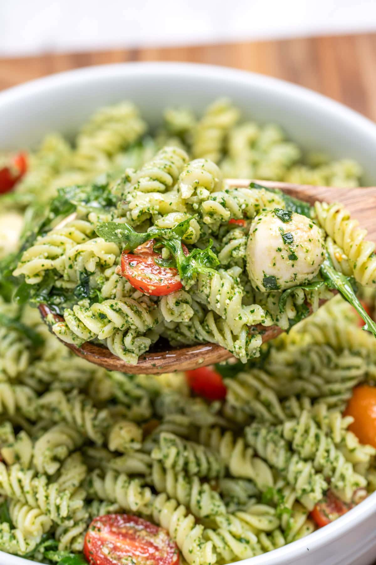 Pesto pasta salad with mozzarella and cherry tomatoes in a serving bowl resting on a cutting board, with a wooden spoon lifting some up.