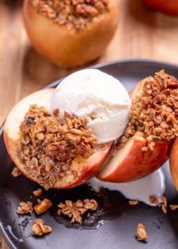 A plate with a baked apple with oat stuffing cut in half and topped with a scoop of vanilla ice cream, with another stuffed apple in the background.