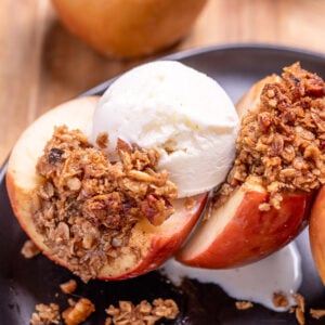 A plate with a baked apple with oat stuffing cut in half and topped with a scoop of vanilla ice cream, with another stuffed apple in the background.