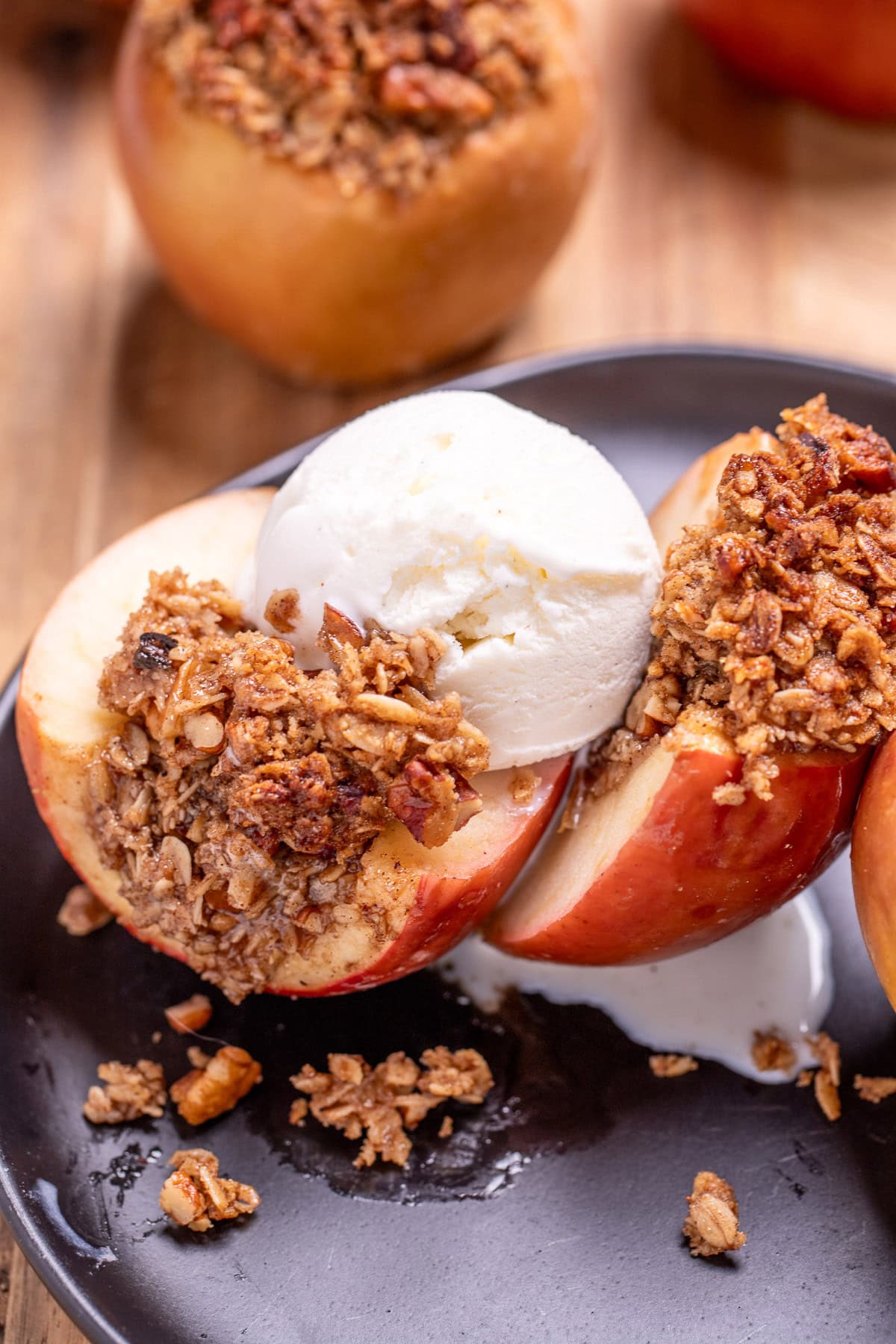 A plate with a baked apple with oat stuffing cut in half and topped with a scoop of vanilla ice cream, with another stuffed apple in the background.