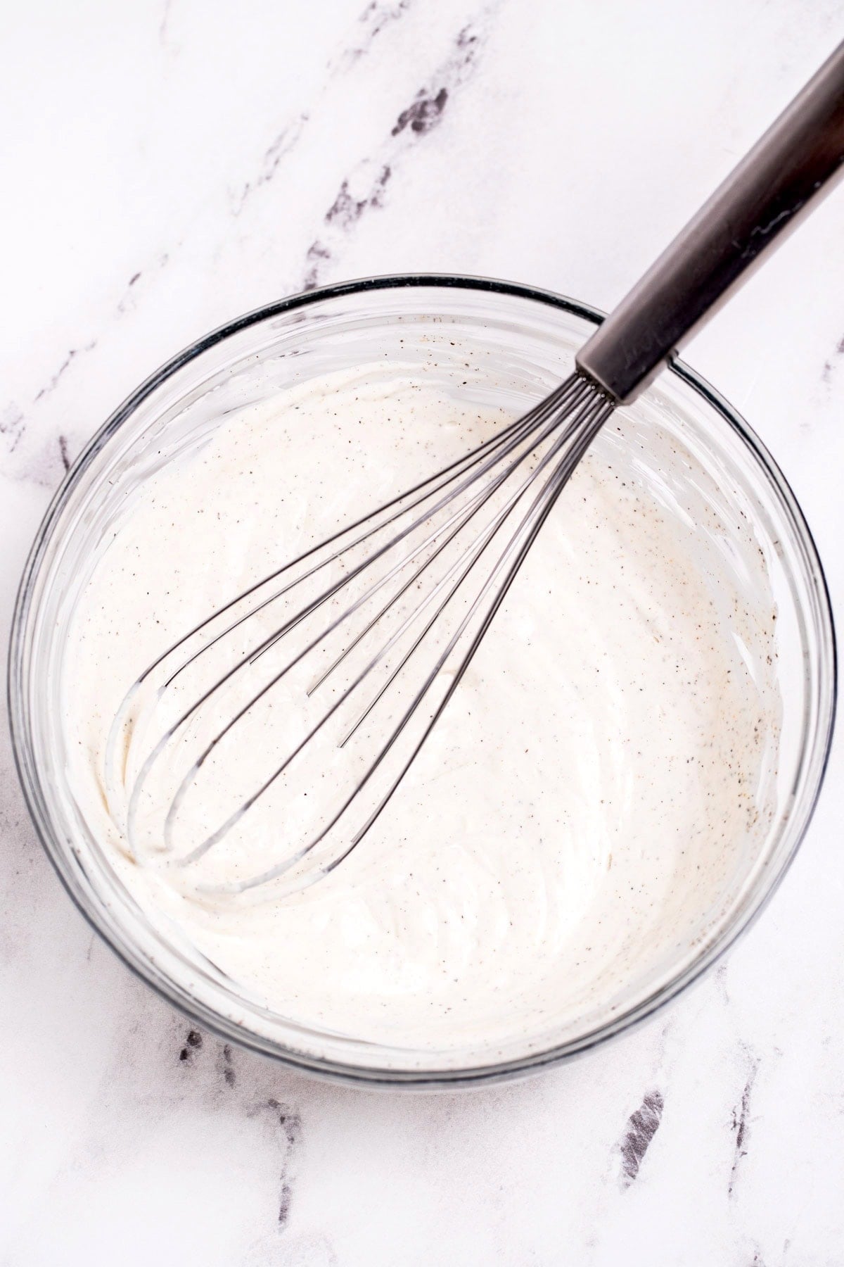 A glass bowl with dill salad dressing on a table, with a whisk resting in the bowl.