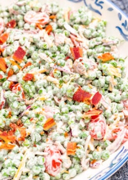 A serving bowl on a tea towel, full of green pea salad topped with crispy bacon.