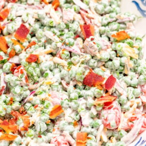 A serving bowl on a tea towel, full of green pea salad topped with crispy bacon.
