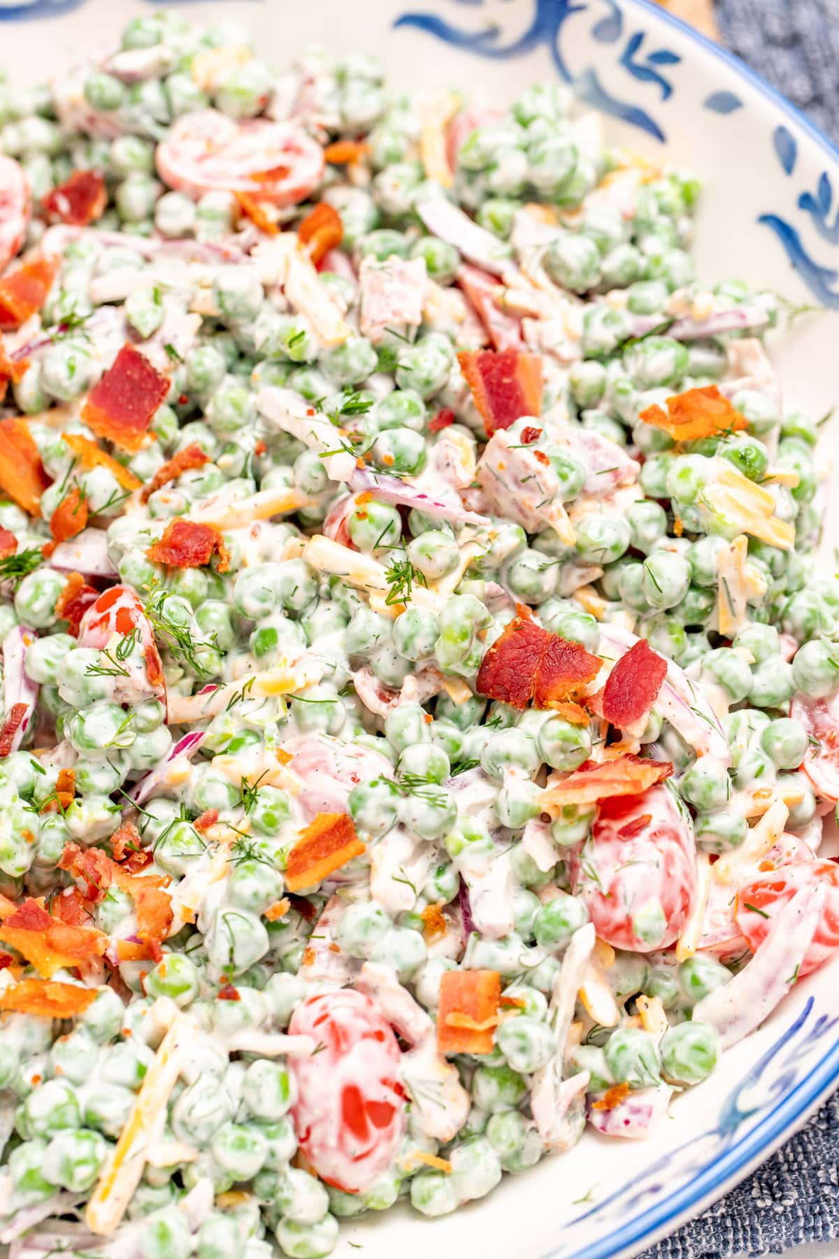 A serving bowl on a tea towel, full of green pea salad topped with crispy bacon.