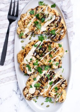 Cauliflower steaks on a serving dish, topped with drizzled tahini dressing, fresh parsley, and pumpkin seeds. On a table with a tea towel and serving fork.