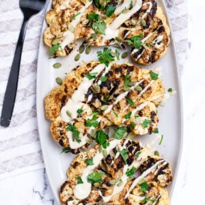 Cauliflower steaks on a serving dish, topped with drizzled tahini dressing, fresh parsley, and pumpkin seeds. On a table with a tea towel and serving fork.
