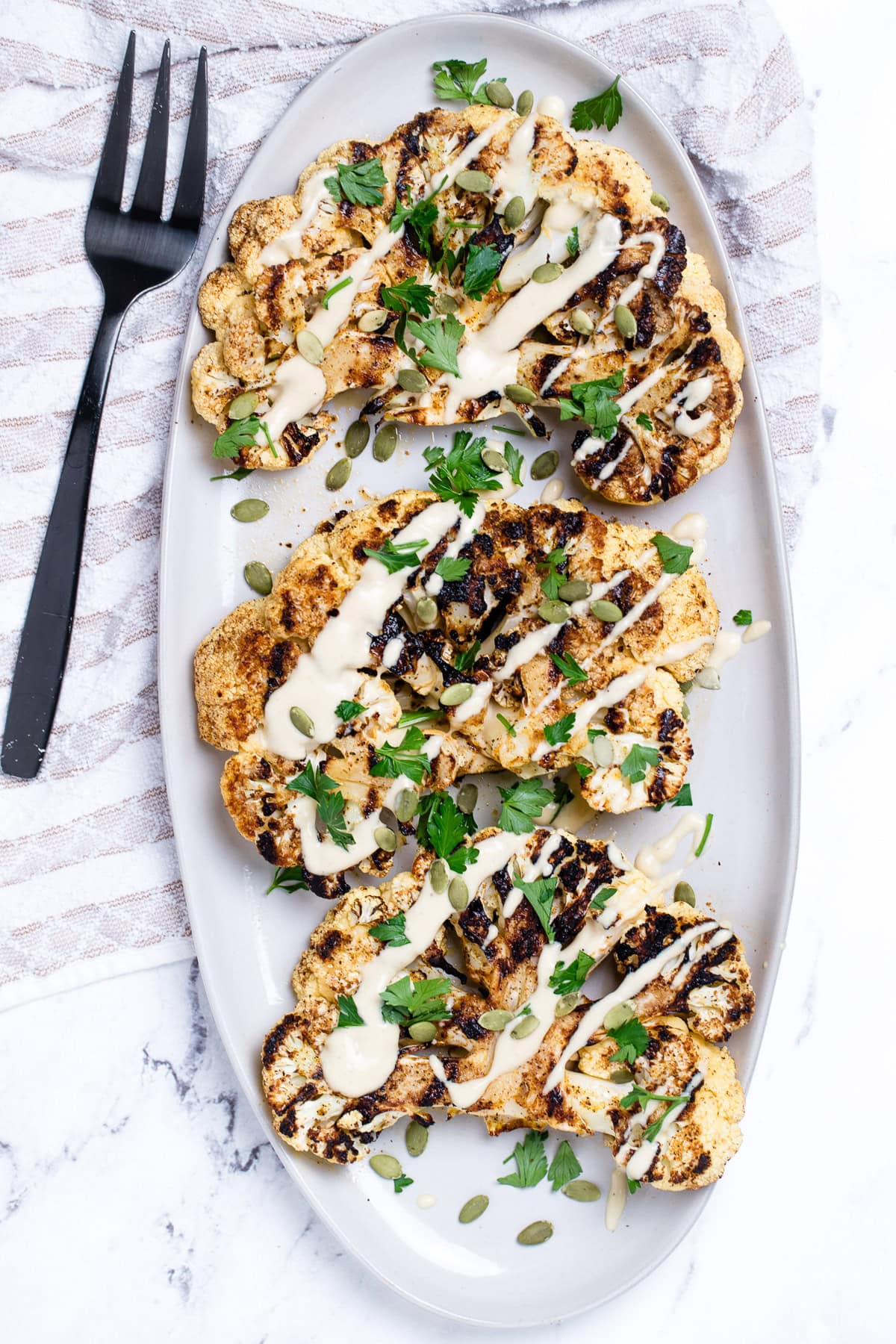 Cauliflower steaks on a serving dish, topped with drizzled tahini dressing, fresh parsley, and pumpkin seeds. On a table with a tea towel and serving fork.