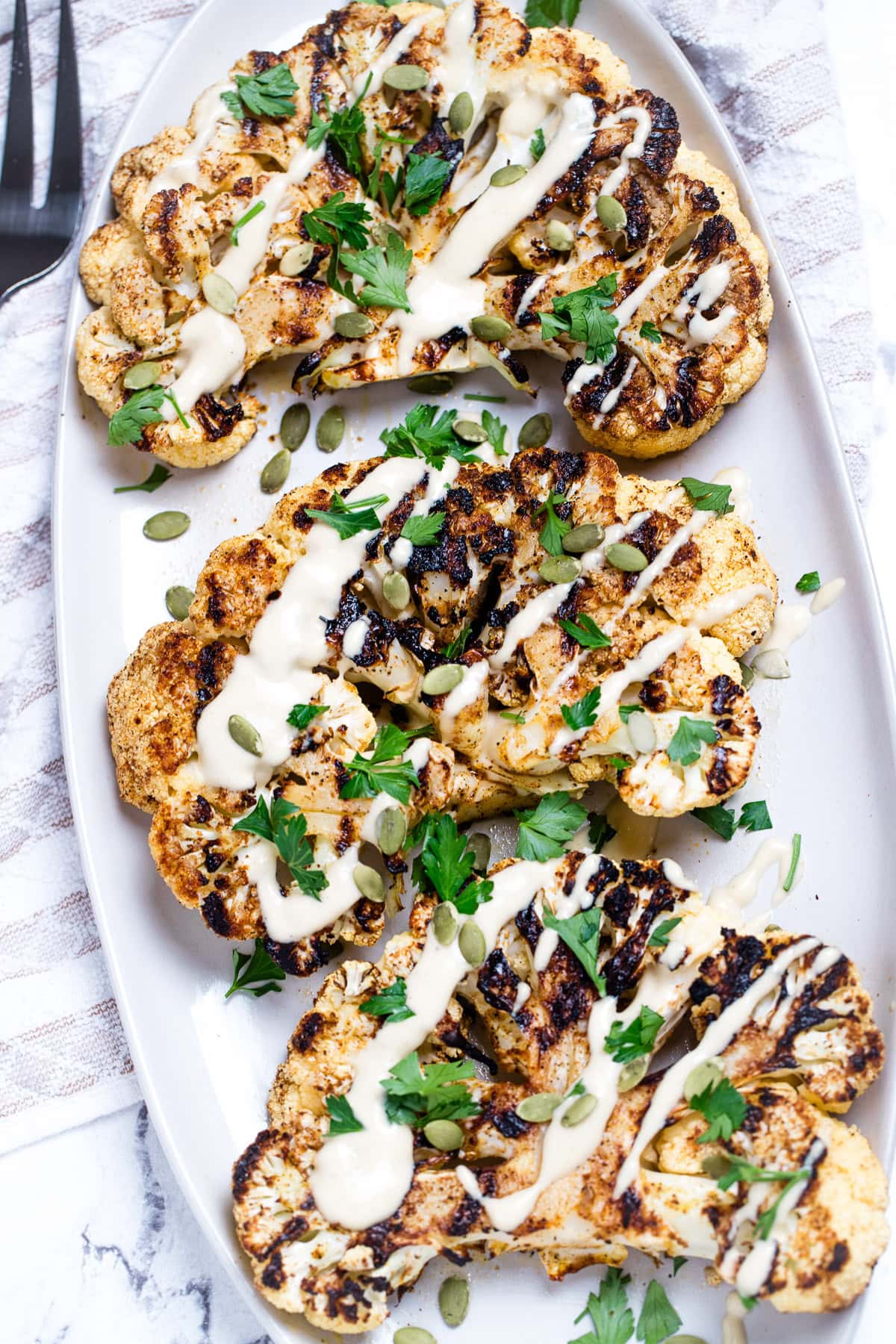 Cauliflower steaks on a serving dish, topped with drizzled tahini dressing, fresh parsley, and pumpkin seeds. On a table with a tea towel and serving fork.