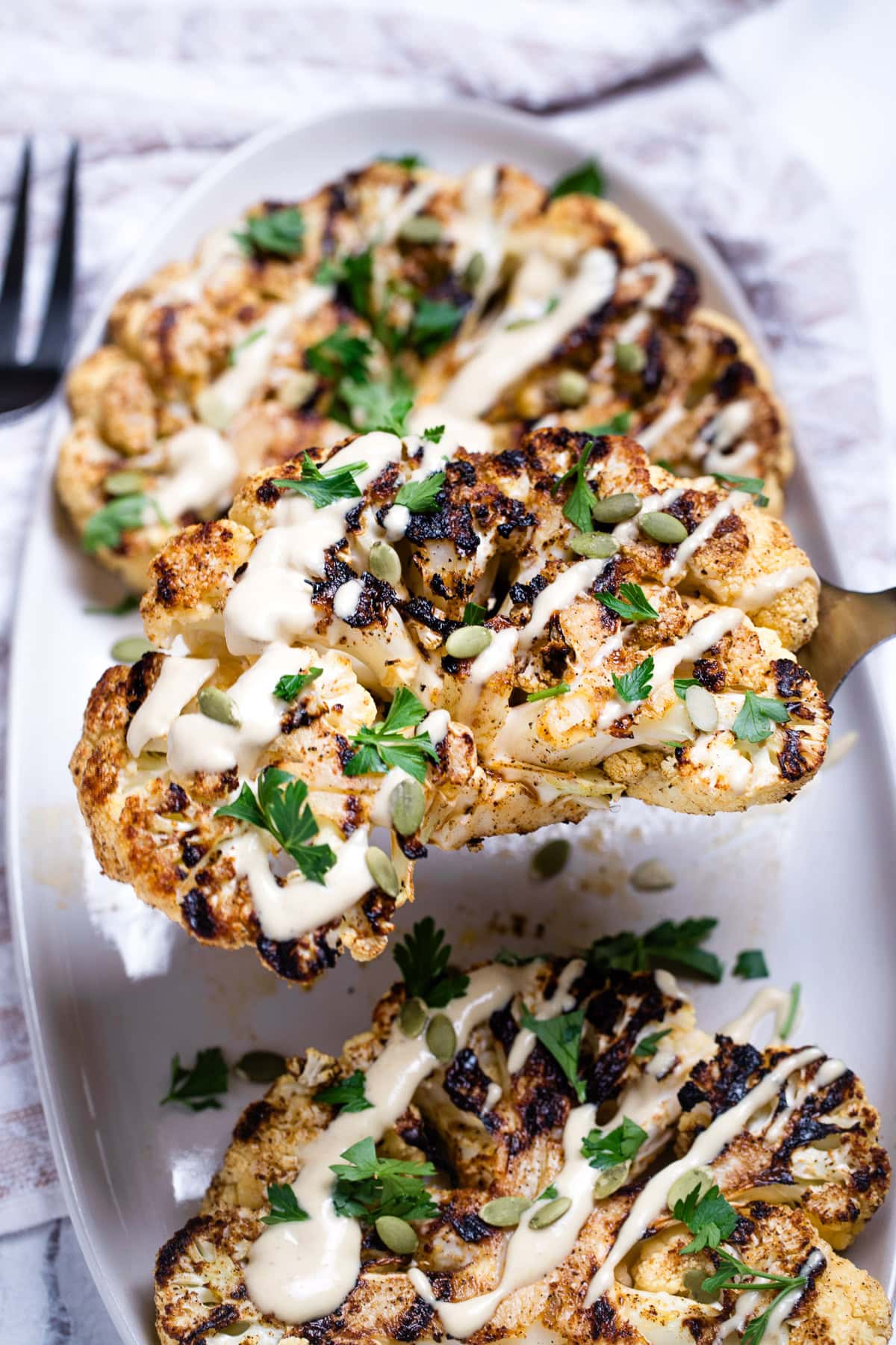 Cauliflower steaks on a serving dish, topped with drizzled tahini dressing, fresh parsley, and pumpkin seeds, with a spatula lifting one up.