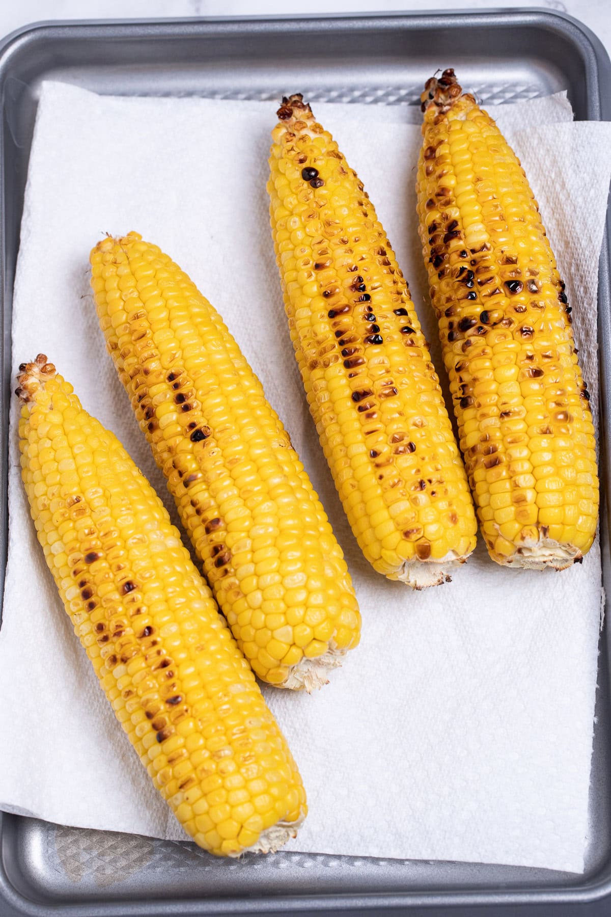 Four ears of corn after being grilled, on a sheet pan lined with paper towels.