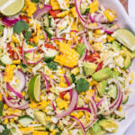A serving bowl full of summer corn salad with corn, avocado, red onion, cilantro, cabbage, and lime wedges, with a wooden spoon and cilantro behind the bowl.
