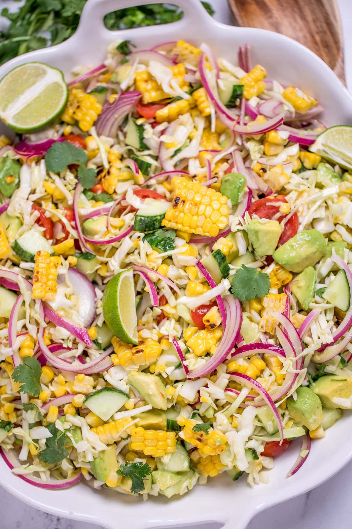 A serving bowl full of summer corn salad with corn, avocado, red onion, cilantro, cabbage, and lime wedges, with a wooden spoon and cilantro behind the bowl.