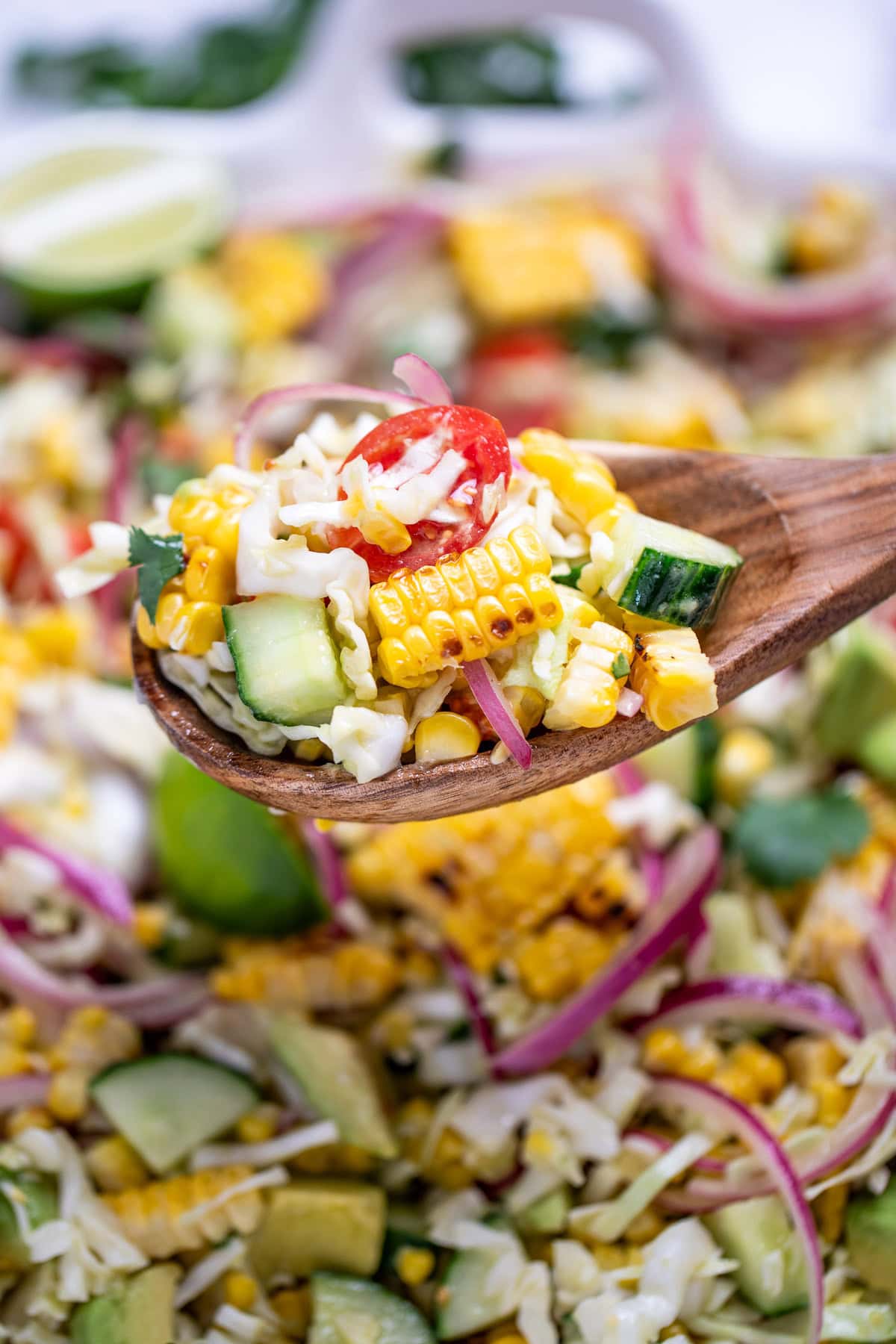 A serving bowl full of summer corn salad with corn, avocado, red onion, cilantro, cabbage, and lime wedges, with a wooden spoon and cilantro behind the bowl, and a spoon lifting some up.