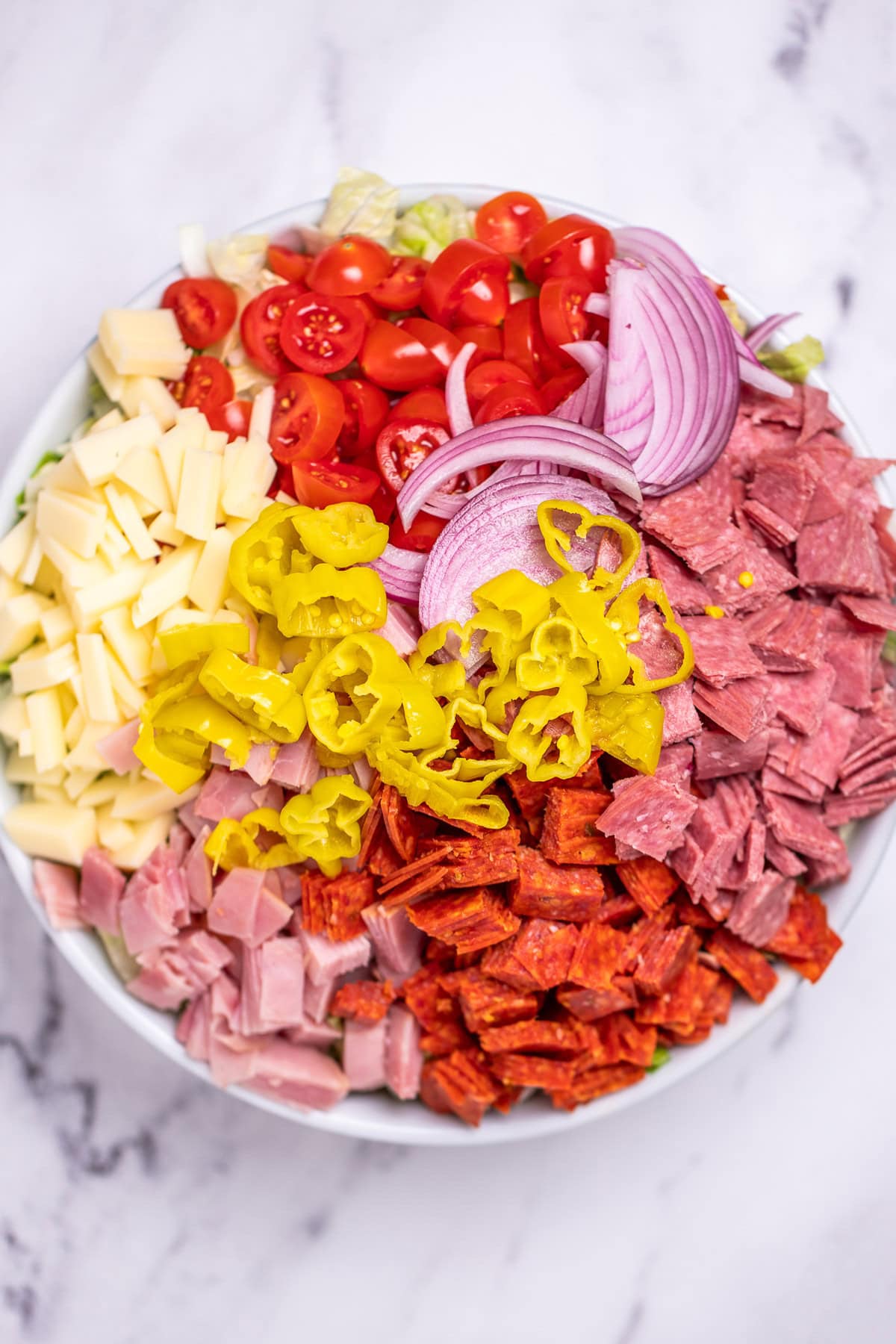 A bowl of salad with sections of ingredients on top, deli meat, provolone cheese, red onion, tomato, and pepperoncini.