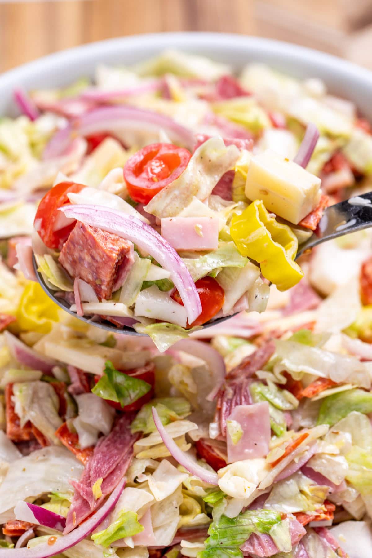 A spoon lifting Italian grinder salad out of a bowl, with lettuce, deli meat and cheese, red onion, and tomato in a creamy dressing.