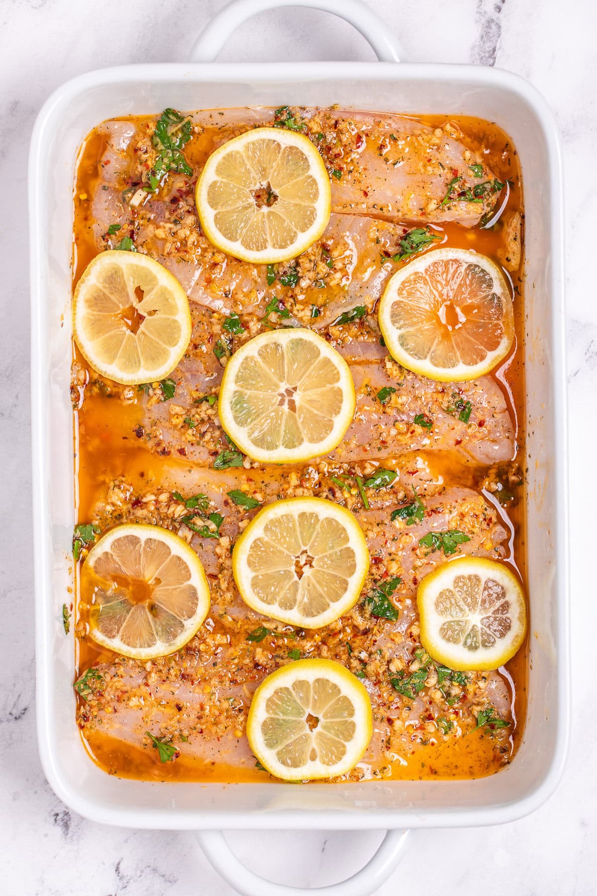 A baking dish with raw tilapia fillets topped with lemon butter sauce and lemon slices.
