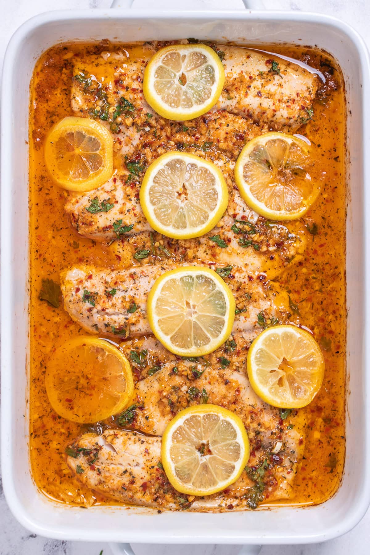 A baking dish with cooked tilapia fillets topped with garlic lemon butter sauce and lemon slices.