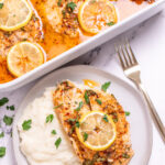 A tilapia fillet with a lemon slice on top of mashed potatoes on a plate, on a table in front of a baking dish with more tilapia in it.