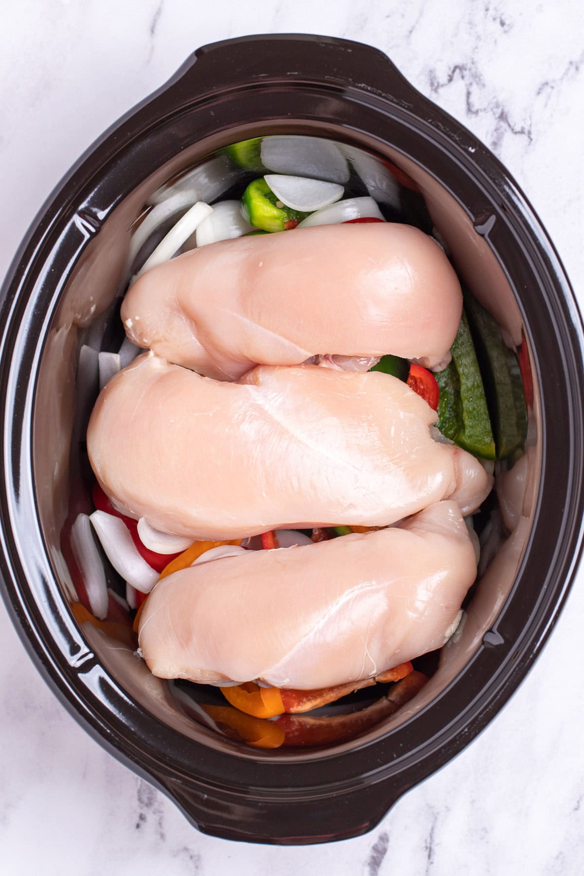 A slow cooker with raw chicken breast resting on top of sliced peppers and onions.