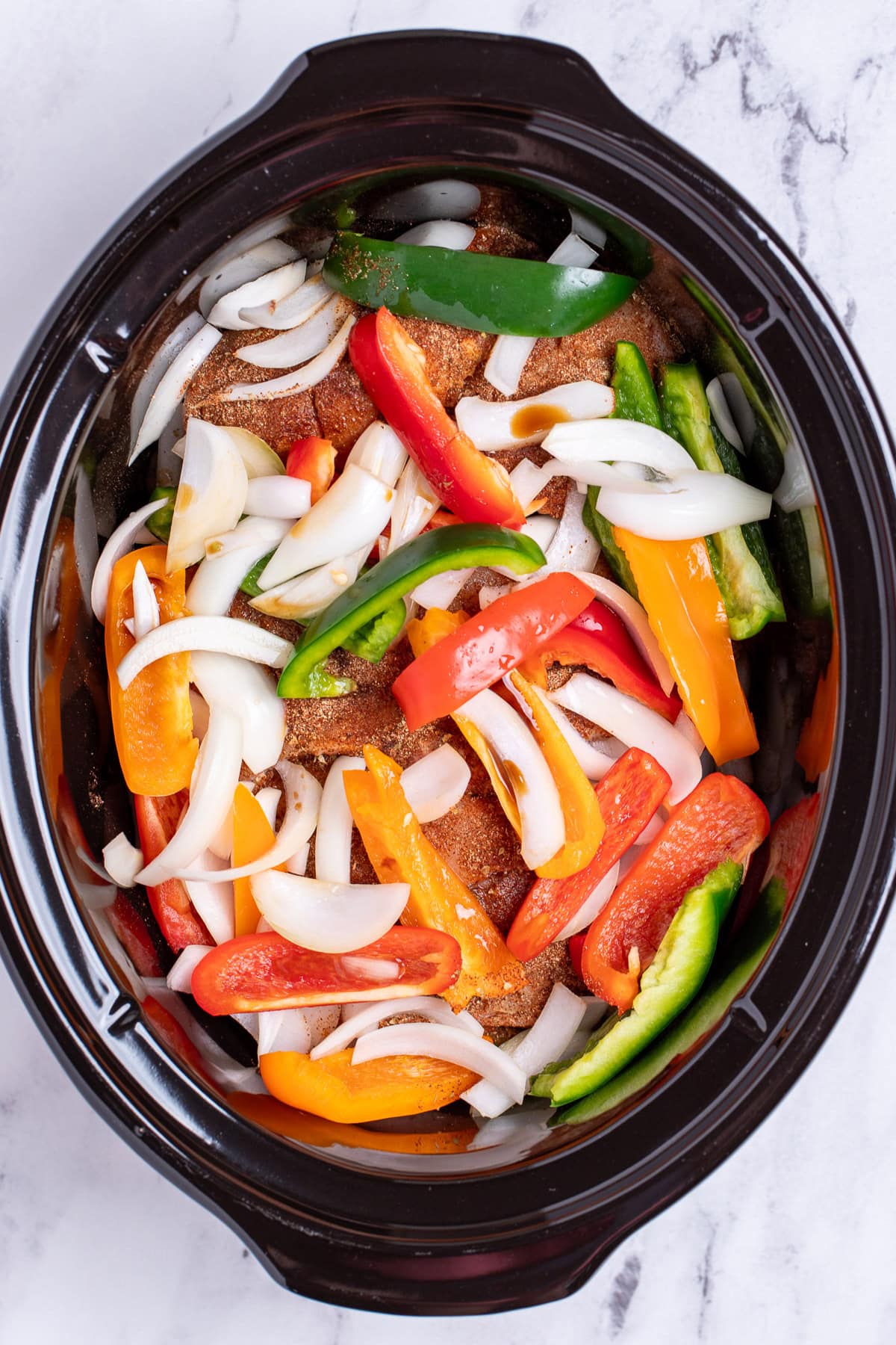 A slow cooker with peppers and onion on top of seasoned chicken breast before being cooked.