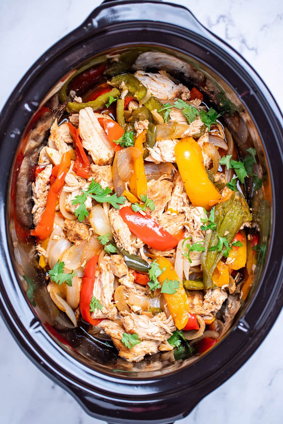 A slow cooker with pieces of seasoned chicken, bell peppers, and onion topped with fresh cilantro.