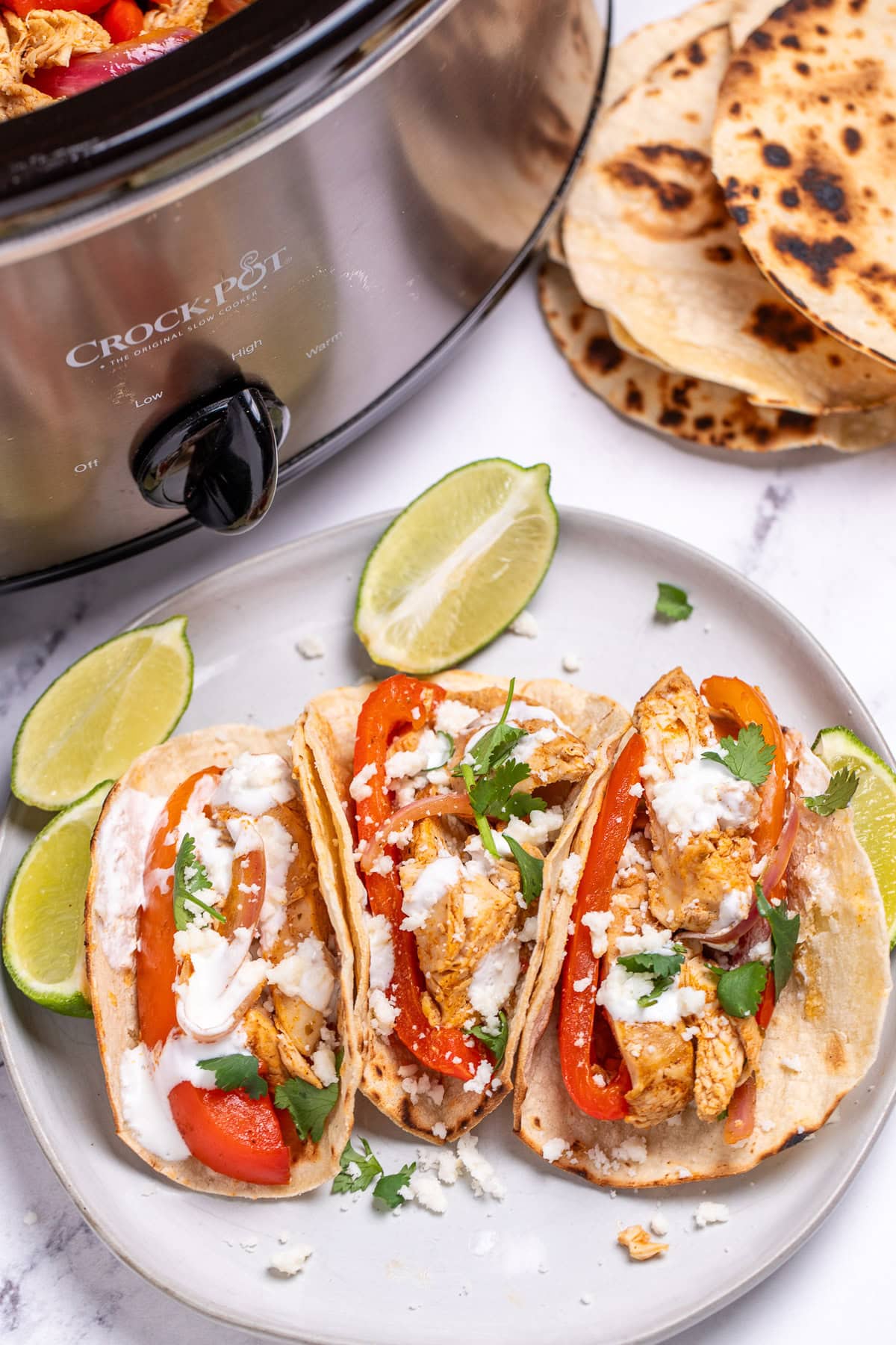 A plate with three corn tortillas with chicken fajitas inside, topped with queso fresco, sour cream, and fresh cilantro, next to lime wedges and a slow cooker, with more tortillas in the background.