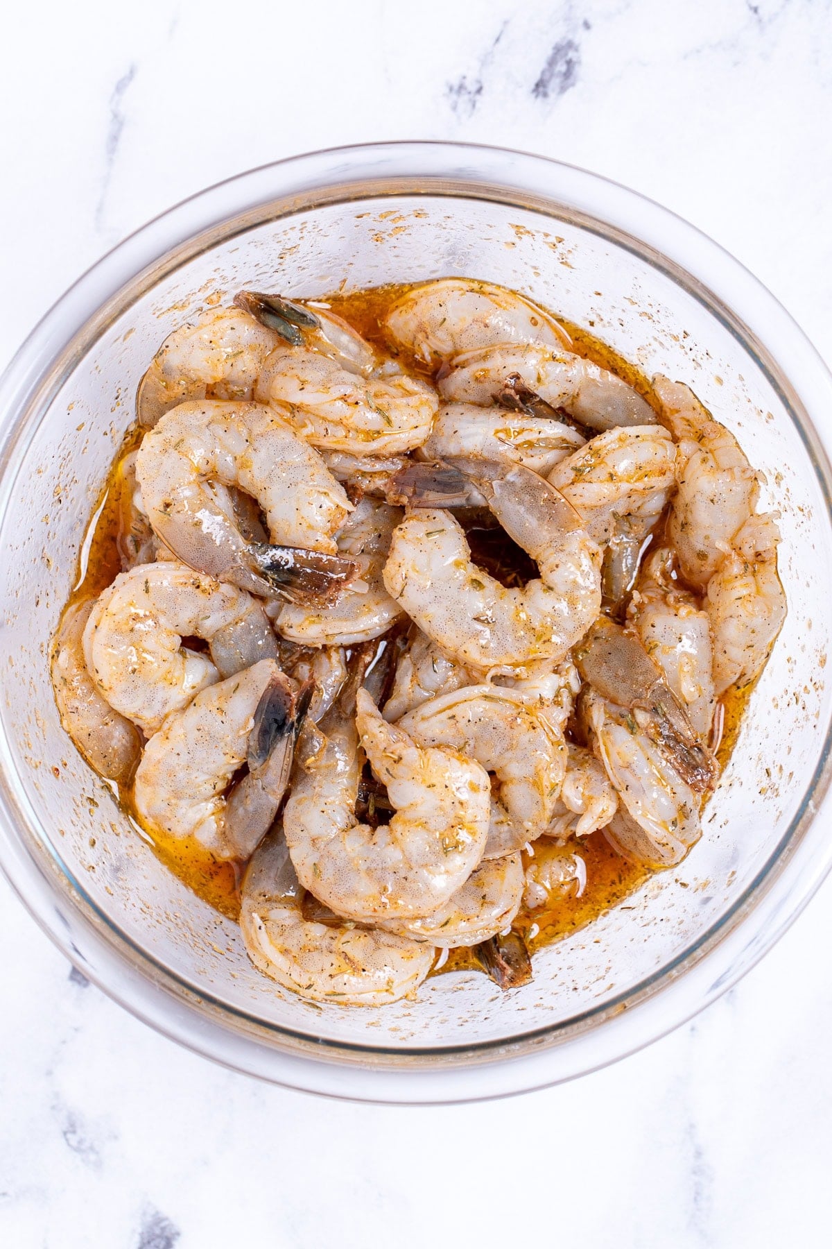 A glass bowl on a table, full of raw shrimp coated in a marinade.