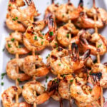 A plate on a table with grilled shrimp skewers topped with green onion, and a hand holding one up.