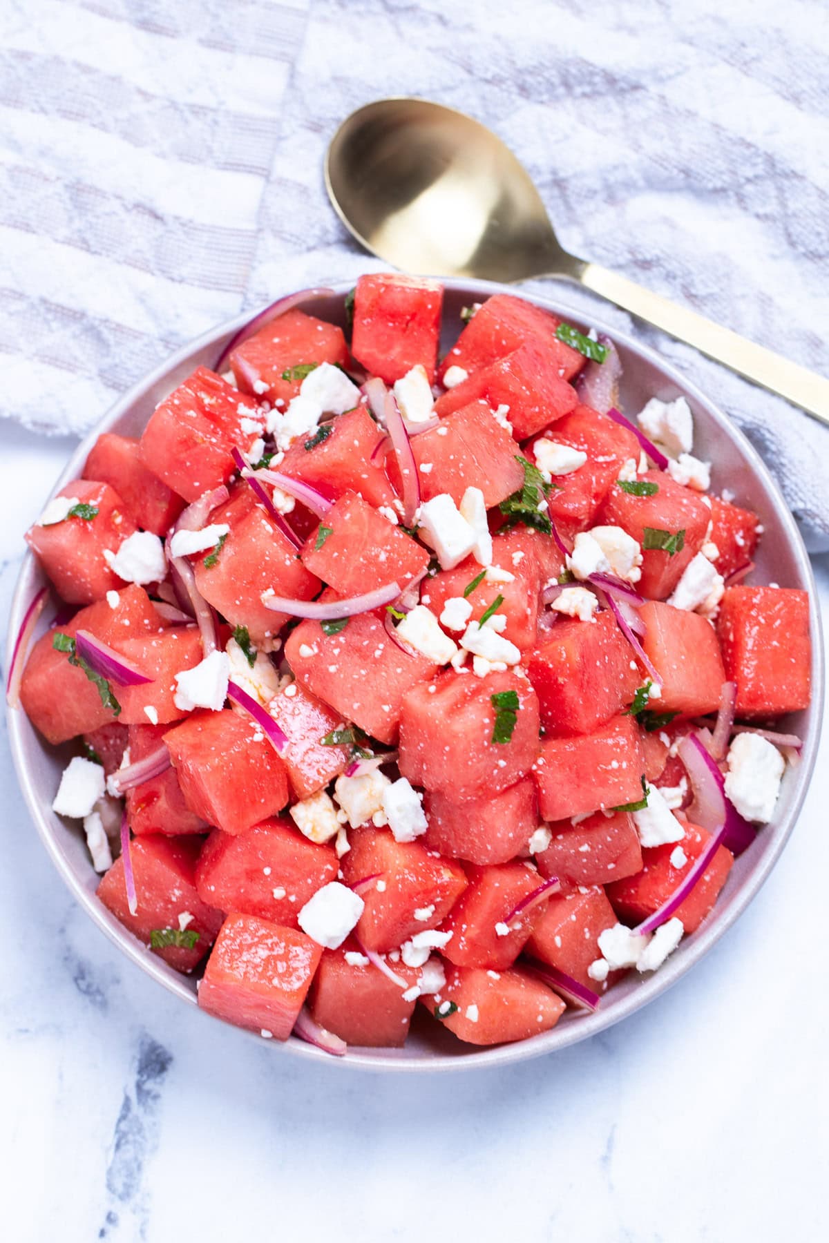 Watermelon feta salad in a bowl on a table next to a serving spoon and a tea towel.