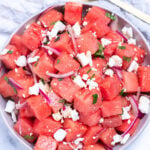 Watermelon feta salad in a bowl on a table next to a serving spoon and a tea towel.