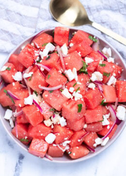 Watermelon feta salad in a bowl on a table next to a serving spoon and a tea towel.