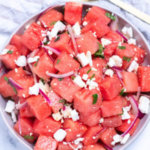 Watermelon feta salad in a bowl on a table next to a serving spoon and a tea towel.